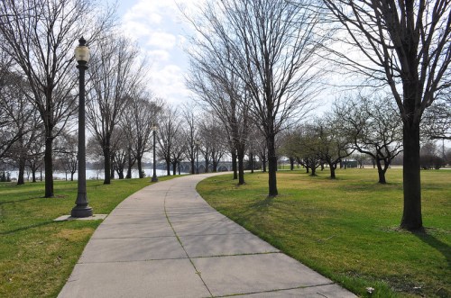 A winding path through a park lined with bare trees, leading to a body of water under a partly cloudy sky.
