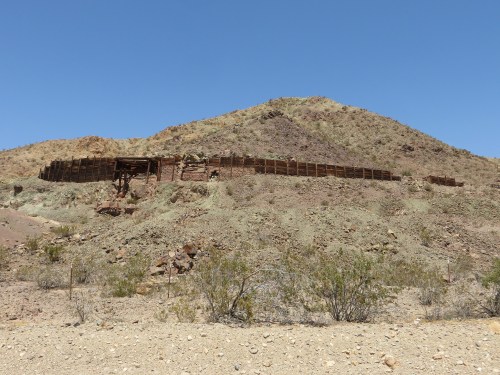 A weathered stone structure on a hillside, surrounded by arid terrain and sparse vegetation under a clear blue sky.