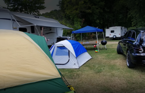 A campsite with tents, a blue canopy, a grill, and parked vehicles in a grassy area surrounded by trees.