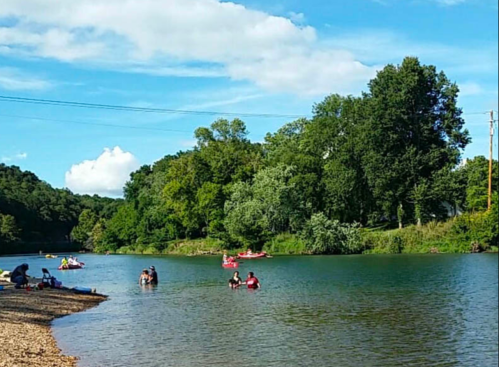 A serene river scene with people kayaking and swimming, surrounded by lush greenery and a clear blue sky.