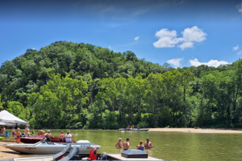 A sunny riverside scene with people relaxing on a dock and boats, surrounded by lush green trees and a blue sky.