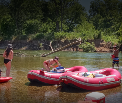 People enjoying a sunny day on the water, using red inflatable rafts and surrounded by greenery.