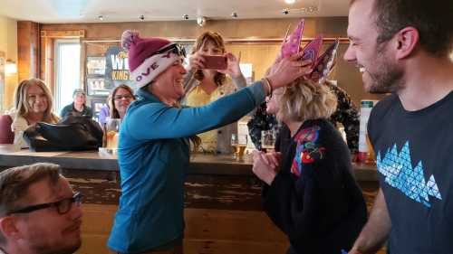 A woman places a colorful crown on another woman's head while friends celebrate in a lively bar setting.