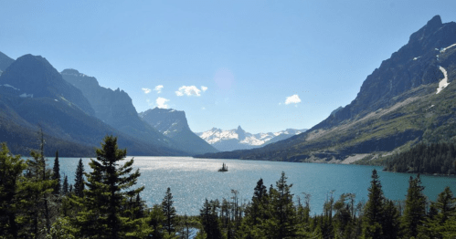 A serene lake surrounded by mountains and lush trees under a clear blue sky. Snow-capped peaks in the distance.