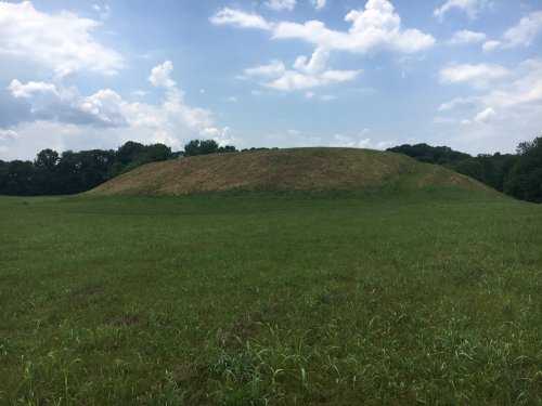 A grassy mound rises in a field under a partly cloudy sky, surrounded by trees in the background.