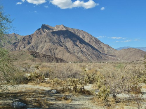 A rugged mountain rises above a dry, desert landscape with sparse vegetation under a clear blue sky.