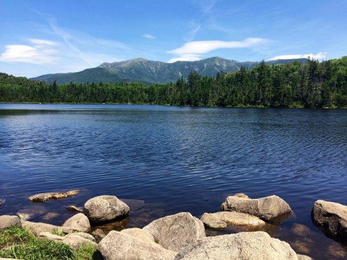 A serene lake surrounded by lush greenery and mountains under a clear blue sky, with rocks in the foreground.