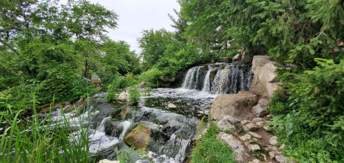 A serene waterfall cascades over rocks, surrounded by lush greenery and trees in a peaceful natural setting.