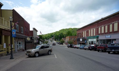 A quiet small-town street lined with shops and parked cars, surrounded by green hills under a cloudy sky.
