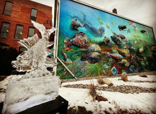 An ice sculpture in front of a vibrant mural depicting colorful sea creatures on a brick building.