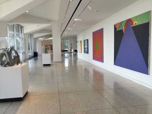 A spacious art gallery featuring colorful abstract paintings and sculptures displayed on white walls and pedestals.