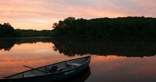 A calm lake at sunset, with a canoe anchored near the shore and trees reflecting in the water.