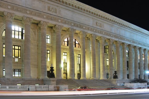 A grand building with tall columns and illuminated windows, captured at night with light trails from passing cars.