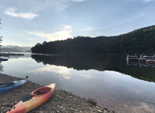 A serene lake at sunrise, with kayaks on the shore and mountains reflected in the calm water.