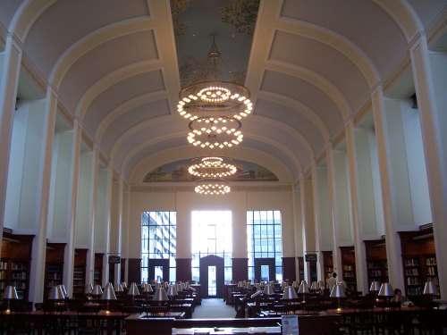 A spacious library interior with high ceilings, elegant chandeliers, and rows of bookshelves. Natural light floods in.