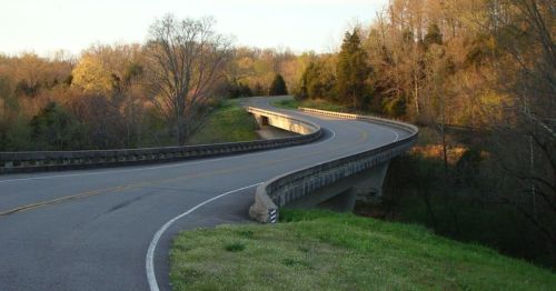 A winding road curves over a bridge, surrounded by trees and greenery in a serene landscape.