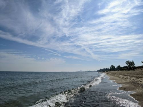 A serene beach scene with gentle waves, sandy shore, and a clear blue sky with wispy clouds.