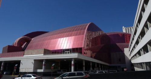 A large, modern building with a shiny, red exterior under a clear blue sky, surrounded by parked cars.
