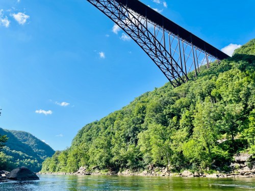 A scenic view of a river surrounded by lush green hills, with a large bridge arching overhead under a clear blue sky.