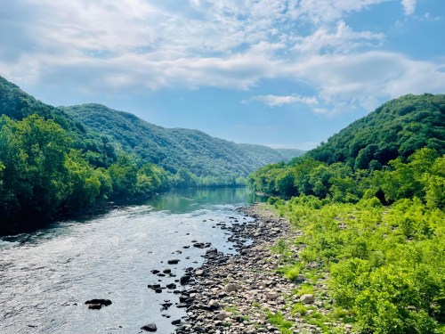 A serene river flows between lush green hills under a bright blue sky with scattered clouds.