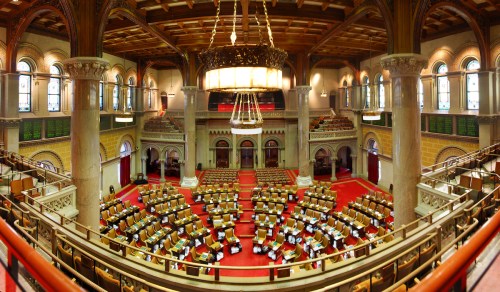 A panoramic view of a grand legislative chamber with rows of seats, ornate architecture, and large windows.