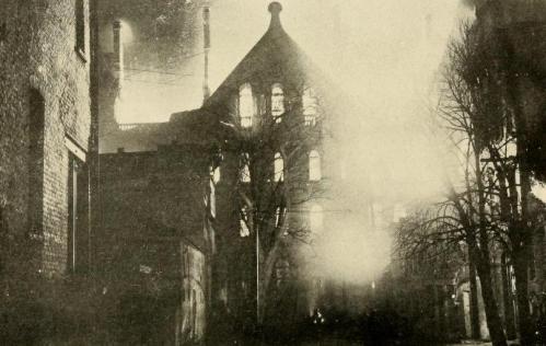 A historic black-and-white photo of a building shrouded in smoke, with trees and nearby structures visible.
