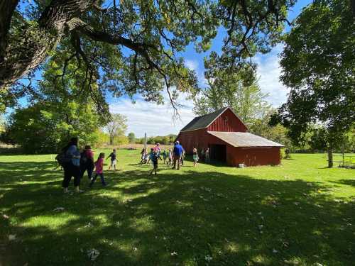 A group of people walks towards a red barn on a sunny day, surrounded by green grass and trees.