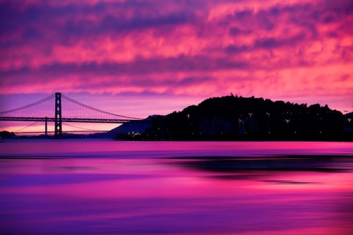 A vibrant sunset casts purple hues over a bridge and calm waters, with silhouettes of trees and buildings in the background.