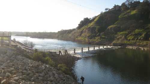 A serene river scene with a person fishing, surrounded by lush hills and a wooden bridge in the background.