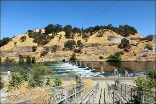 A river with a small dam, surrounded by dry hills and greenery, under a clear blue sky. Steps lead down to the water.