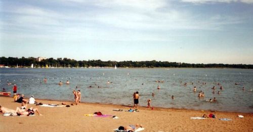A sandy beach with people sunbathing and swimming in a calm lake under a clear blue sky.