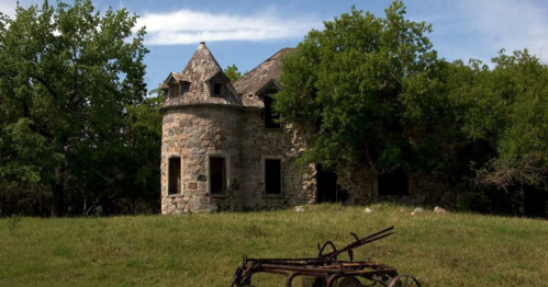 Abandoned stone house with a turret, surrounded by trees and a grassy field, featuring an old plow in the foreground.