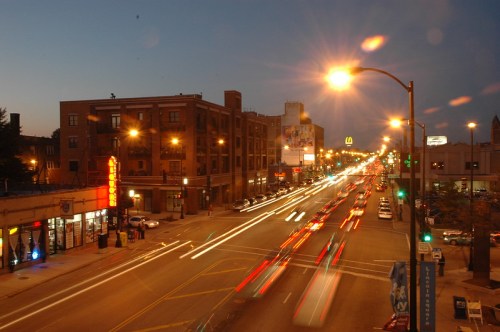 A busy city street at dusk, with light trails from moving cars and illuminated buildings lining the road.