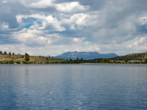 A serene lake reflects cloudy skies and distant mountains, surrounded by lush greenery and rolling hills.