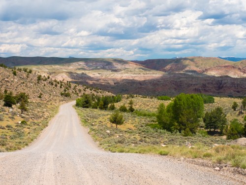 A winding gravel road leads through a rugged landscape of hills and sparse trees under a cloudy sky.