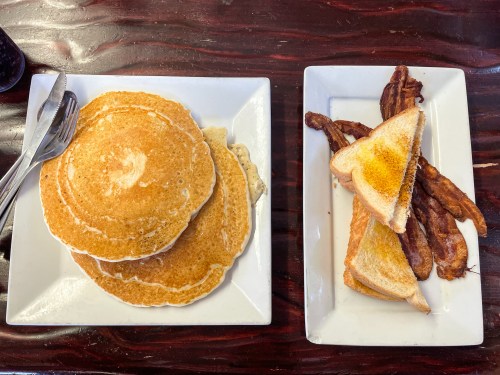 A plate with three fluffy pancakes and another plate with toast and crispy bacon strips.