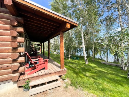 A log cabin porch with red chairs overlooks a grassy area and a serene lake surrounded by trees.