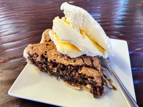 A slice of chocolate pie topped with a scoop of vanilla ice cream on a white plate, with a fork beside it.