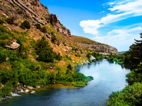 A serene river flows through a lush green landscape, flanked by rocky hills under a bright blue sky.