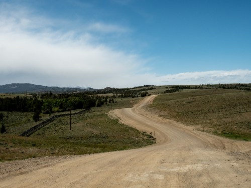 A winding dirt road stretches through a grassy landscape under a blue sky with scattered clouds.