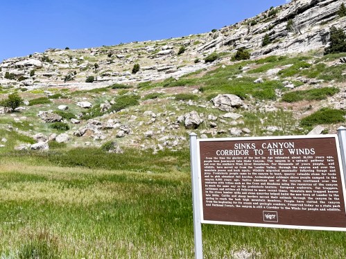 A scenic view of Sinks Canyon with a sign detailing its history, surrounded by green grass and rocky hills.