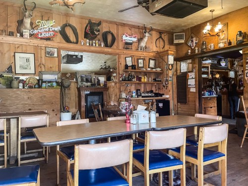 Cozy rustic interior of a bar or restaurant with wooden walls, tables, and various decorations, including antlers and signs.