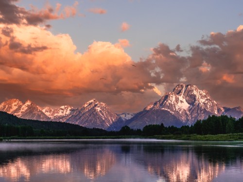 Scenic view of mountains at sunset, reflecting in a calm lake with colorful clouds in the sky.