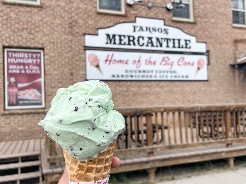 A hand holds a mint chocolate chip ice cream cone in front of a brick building with a sign for "Parson Mercantile."