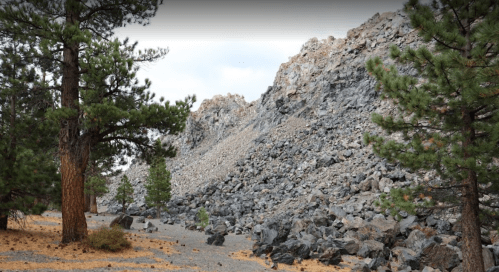 Rocky hillside with scattered boulders, surrounded by pine trees and a gravel path under a cloudy sky.