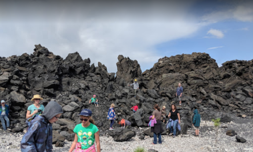 A group of children and adults exploring a rocky, volcanic landscape under a blue sky with scattered clouds.