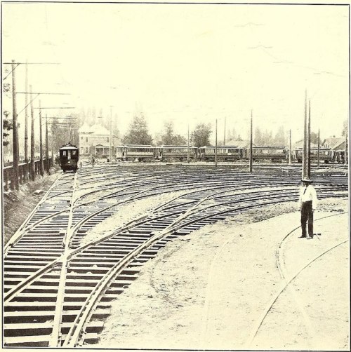 A historic railway scene showing curved tracks and a man standing near the rails, with buildings in the background.