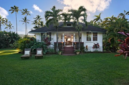 A charming white house surrounded by lush greenery and palm trees, with two lounge chairs in the foreground.