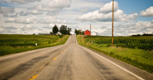 A winding rural road leads towards a red barn and green fields under a cloudy sky.