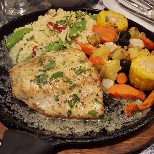 A sizzling platter with grilled fish, couscous, and a colorful mix of roasted vegetables.